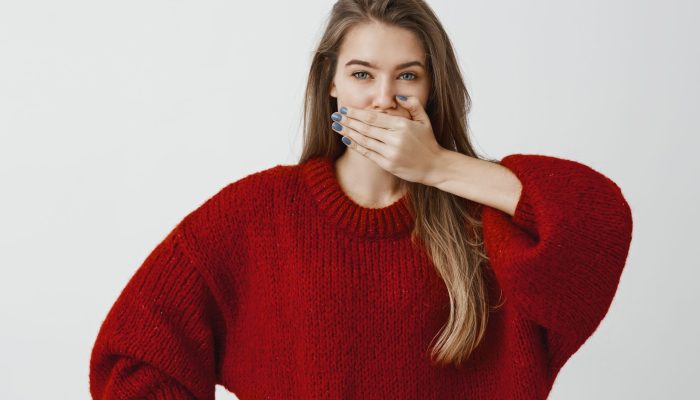 Friend swear not saying word about secret. Portrait of good-looking feminine woman in trendy loose sweater, covering mouth with palm, making promise to keep silence over gray background.