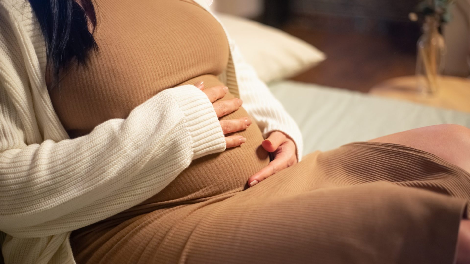 Close-up of pregnant woman sitting on bed. Woman in dress and cardigan touching big belly. Pregnancy, expectation concept