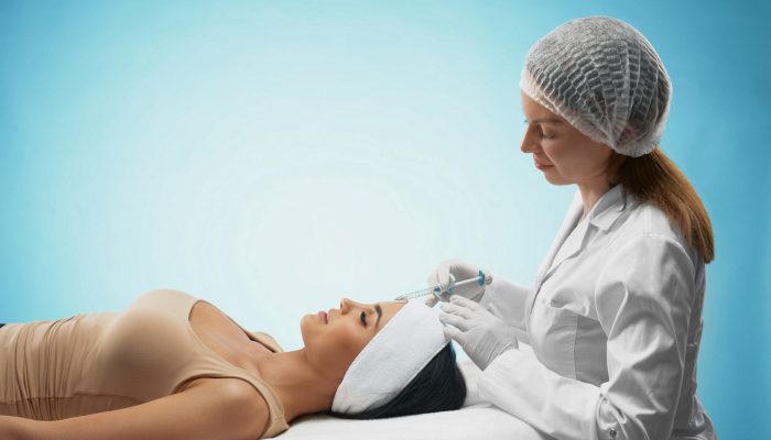 Side view of hyaluronic injection in female forehead. Cosmetologist in coat using syringe with filler while brunette patient with closed eyes, in towel on head lying. Concept of cosmetology, beauty.