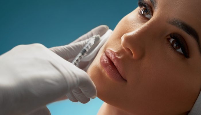 Close up of cosmetic injection in female cheek. Cosmetologist using syringe with special liquid and cotton pad while pretty young patient looking away. Concept of cosmetology, beauty.
