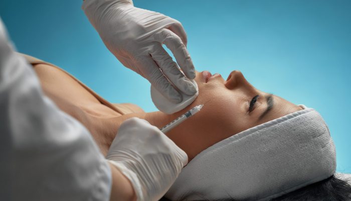 Close up of cosmetic injection in female cheek. Cosmetologist using syringe with special liquid and cotton pad while brunette patient in towel on head lying. Concept of cosmetology, beauty.