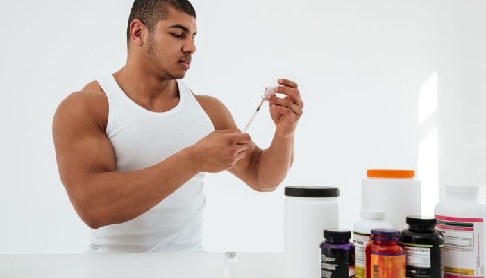 Picture of young sportsman standing over white background holding syringle vitamins standing near sport pills. Looking aside.