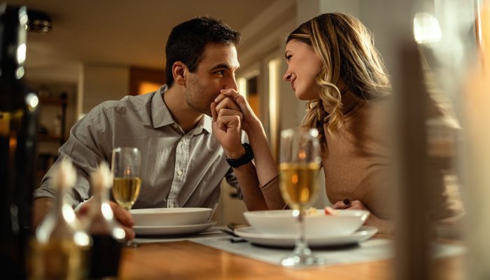 Happy woman enjoying at dining table while being kissed in a hand by her boyfriend.