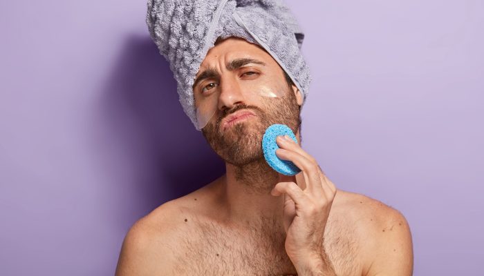 Relaxed serious man with stubble wipes skin on face after shower, holds cosmetic sponge, wrapped in soft towel, stands against purple background. People, masculinity, beauty and wellness concept