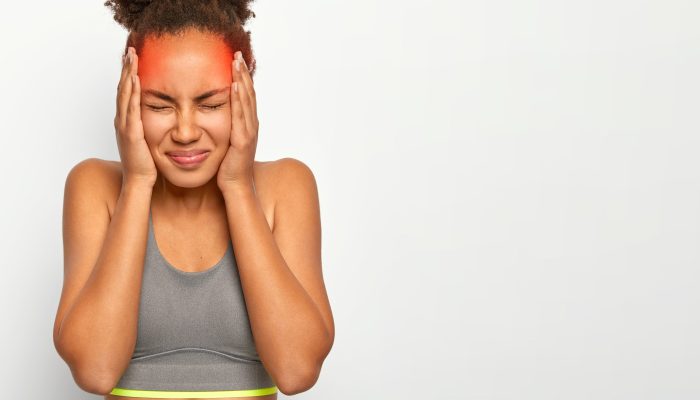 Horizontal shot of dissatisfied curly haired woman touches both temples, suffers from migraine, has marked red location of pain, wears sports bra, closes eyes from bad feelings, isolated on white wall