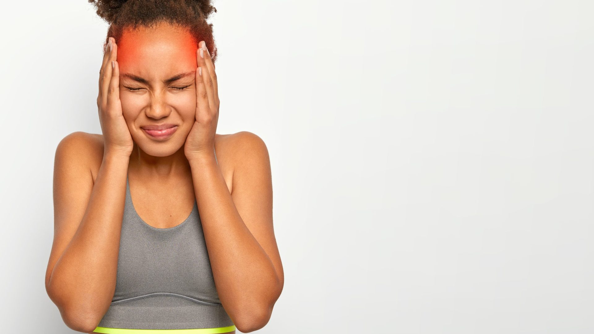 Horizontal shot of dissatisfied curly haired woman touches both temples, suffers from migraine, has marked red location of pain, wears sports bra, closes eyes from bad feelings, isolated on white wall