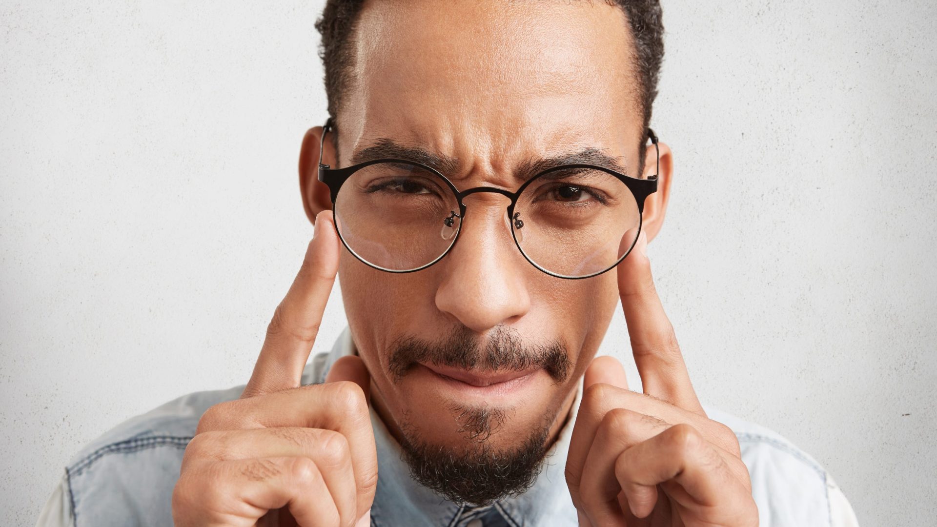Indoor shot of hipster guy in round spectacles keeps fingers on temples, tries to concentrate and find right solution or way out in difficult situation, isolated over white studio background