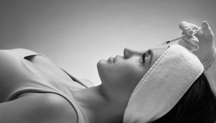 Close up of cosmetic botox injection in female forehead. Cosmetologist using syringe with filler while brunette patient in towel on head lying, isolated on blue. Concept of cosmetology, beauty.