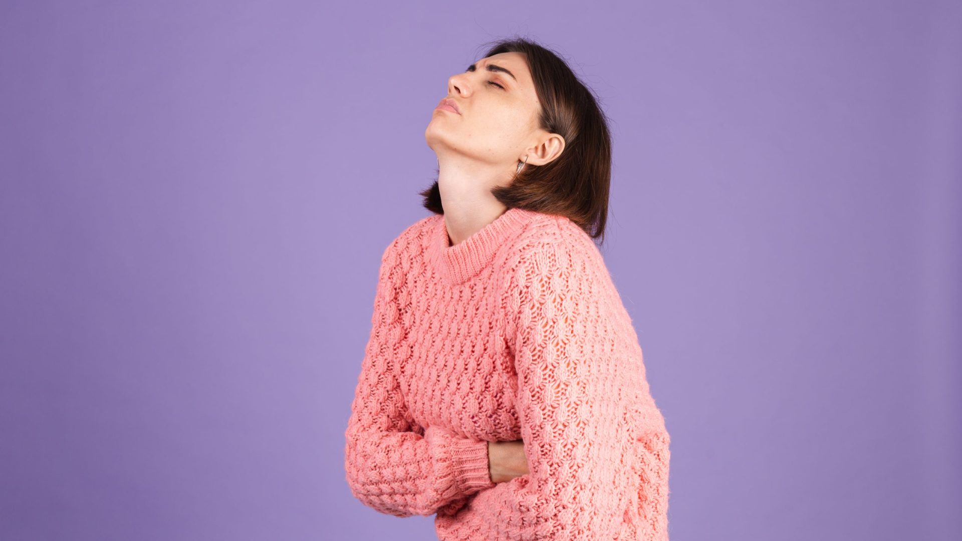 Young brunette in pink sweater isolated on purple background suffering stomach ache with painful grimace, feeling sudden period cramps, gynecology concept