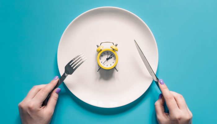 Hands holding knife and fork above alarm clock on a plate on blue background. Intermittent fasting, Ketogenic dieting, weight loss, meal plan, and healthy food concept.