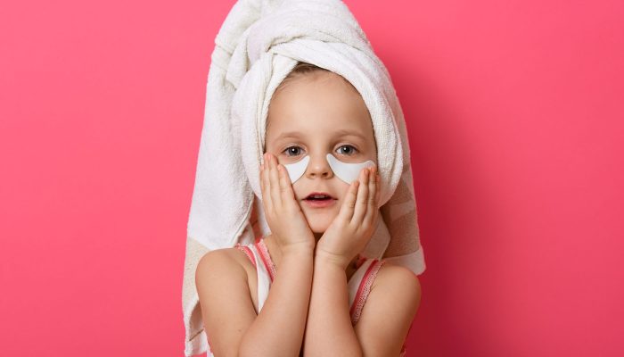 Little cute girl wearing white towel on head, posing with patches under eyes, standing against pink background, charming female kid repeats after mother doing morning procedures.