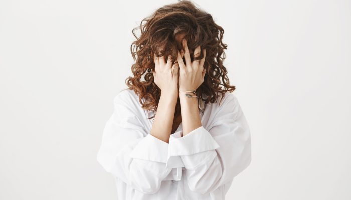 Studio portrait of tired or bothered woman hiding face in her curly hair and holding hands on it, expressing exhaustion or just being childish, standing over gray background. I am not seeing you.
