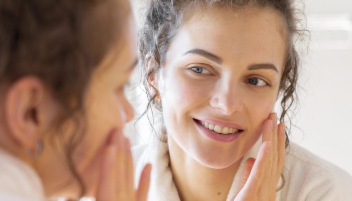 mulher-aplicando-creme-no-rosto-enquanto-se-olha-no-espelho
