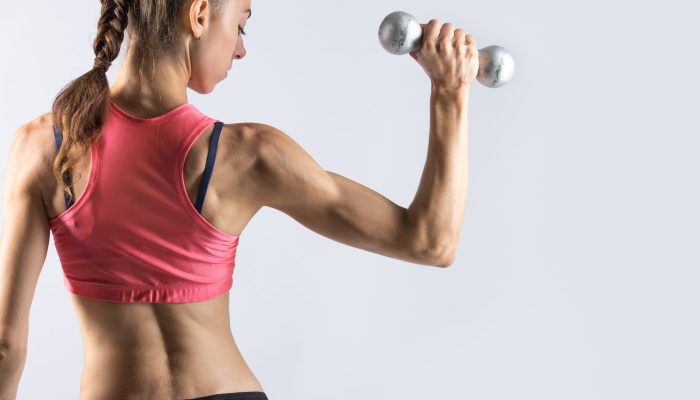 Attractive young fitness person wearing red sportswear top lifting dumbbells. Sporty model girl working out, doing weight training with dumbbells on grey background. Back view. Copy space