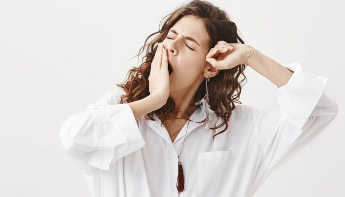 Studio portrait of tired or exhausted young businesswoman yawning and stretching while covering mouth with hand, standing with closed eyes over gray background. Girl have not got enough sleep.