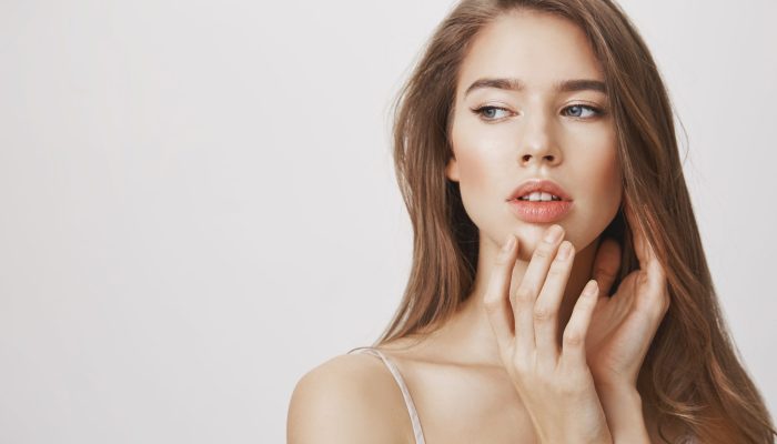 Makeup underline natural beauty. Studio shot of affective romantic caucasian woman with clean skin gently touching chin and looking aside with dreamy expression, standing over gray background. Copy space