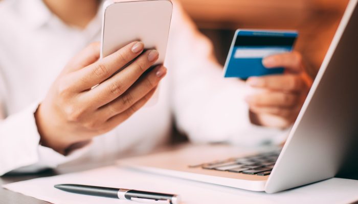 Close-up of female hand holding smartphone. Young businesswoman sitting at laptop in office holding credit card and entering data on her mobile phone