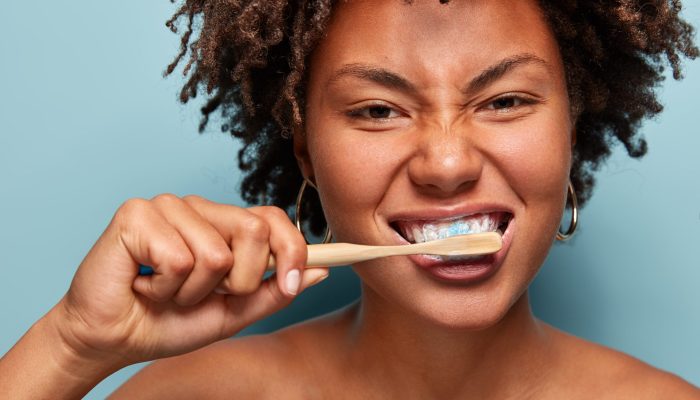 Dental hygiene concept. Self confident mixed race young woman brushes teeth with toothpaste and toothbrush, stands in front of mirror nude, isolated on blue background. Medicine, health care