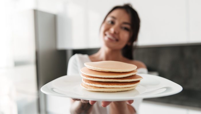 Photo of happy young woman standing at the kitchen in home cooking. Looking camera holding pancakes. Focus on pancakes.