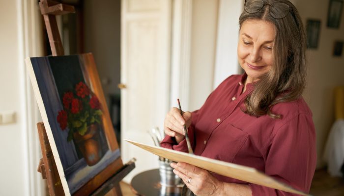 Image of creative skillful middle aged woman artist enjoying her hobby, standing next to easel at home, mixing oil pigments on palette using brush, working on painting of flower pot, smiling