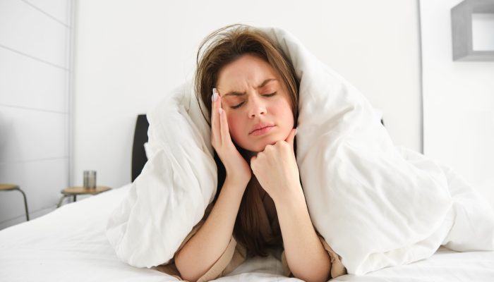 Portrait of woman rolled in blankets in bed, frowning and feeling unwell, has hangover, headache in morning, resting at home.