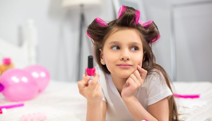 Cute preschooler laying on white blanket holding nail polish. Lovely looking girlie with hair rollers looking at side holding bottle of pink nail polish in her hand.