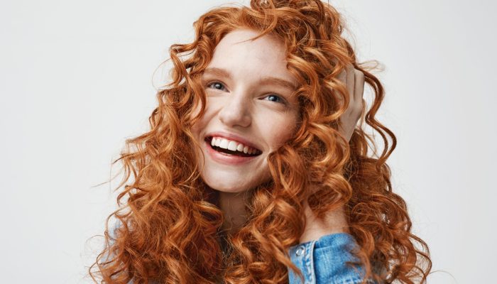 Portrait of cute happy girl laughing touching her curly red hair over white background.