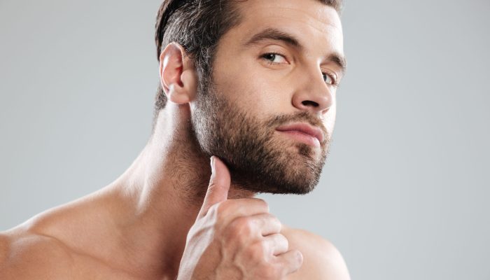 Portrait of a handsome naked bearded man examining his face isolated over white background