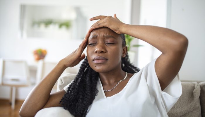 Portrait of a young black girl sitting on the couch at home with a headache and pain. Beautiful woman suffering from chronic daily headaches. Sad woman holding her head because sinus pain