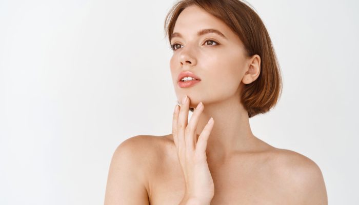 Beauty face. Beautiful young woman looking aside and touching natural healthy skin without make up. Girl with naked shoulders and glowing hydrated face. Skincare concept.