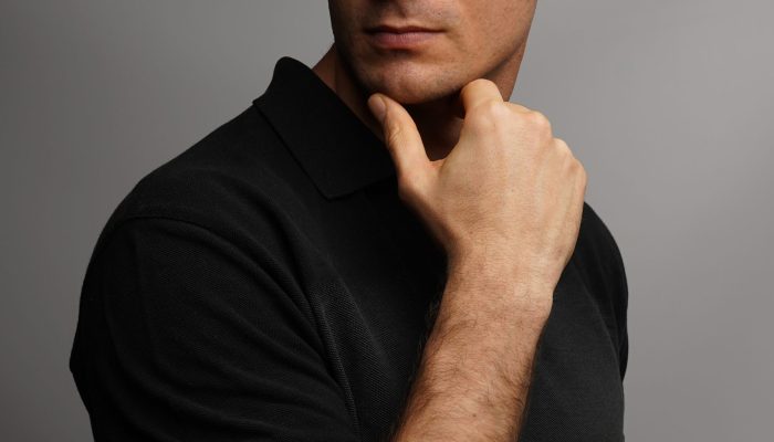The vertical shot of a handsome male holding his chin with an arm against a gray wall