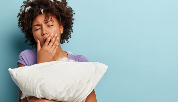 Sleepy tired young Afro American woman covers mouth with hand, yawns after sleep, embraces white pillow, has feathers in head, poses over blue background with blank space for your promotion.