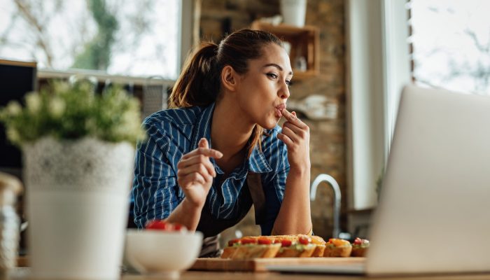 Young happy woman using laptop while preparing bruschetta in the kitchen.